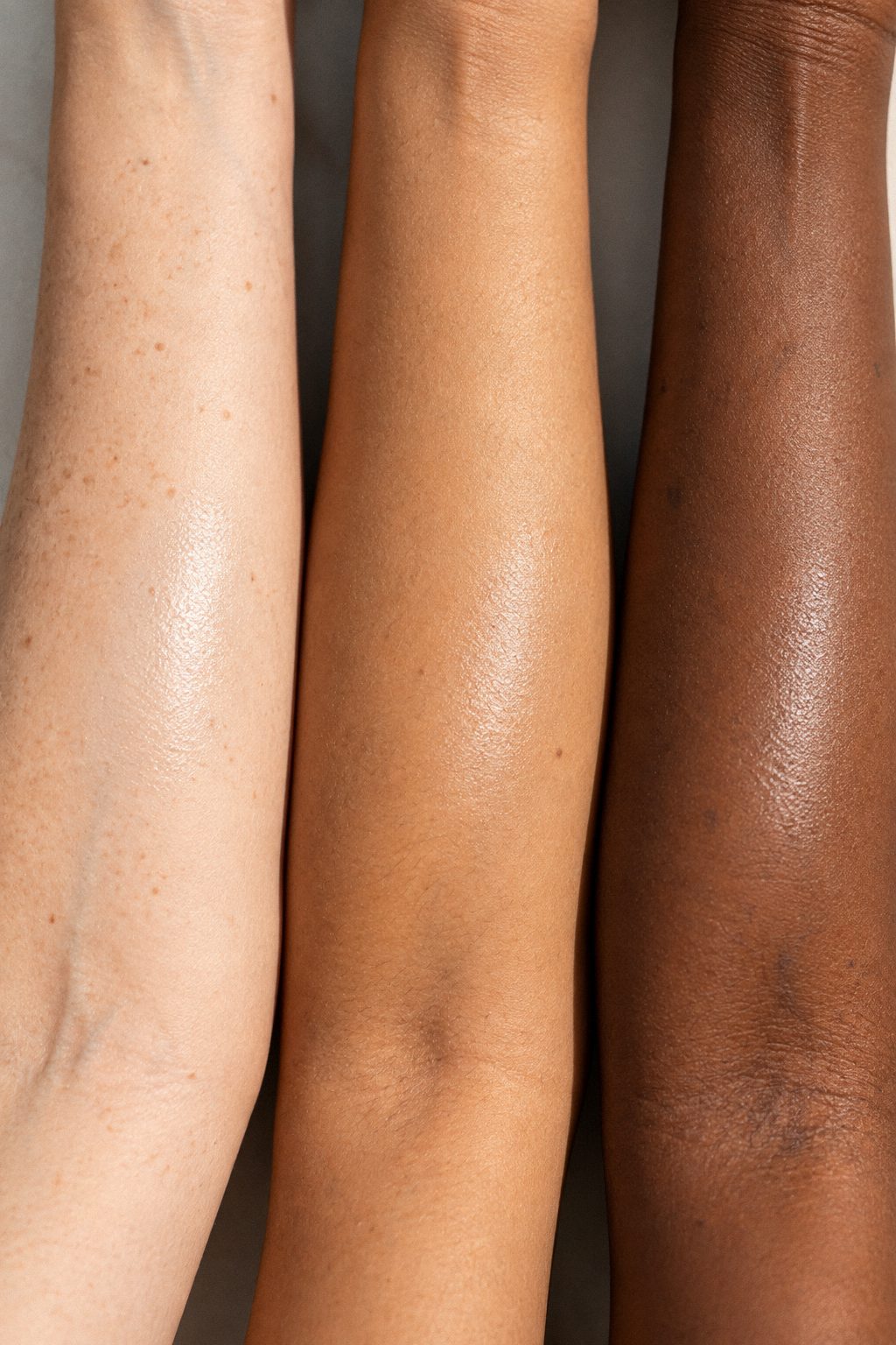 Three forearms side by side, fair, olive and deep brown, each with a swipe of mineral sunscreen blended in until only a soft sheen remains.