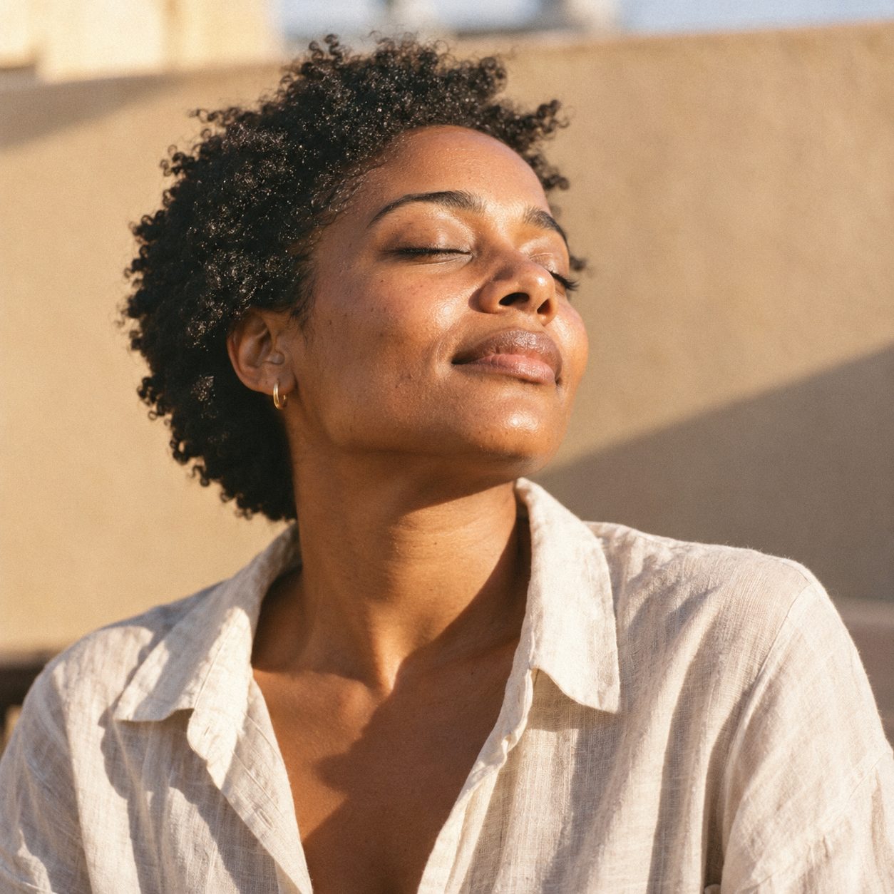 A woman with deep brown skin turns her face, eyes closed, into warm late-afternoon sun.